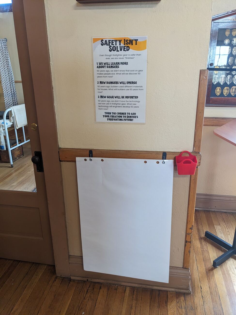 Firefighting in the Future drawing station: a blank flip-chart pad hung by a doorway for visitors to sketch their own answer to Safety Isn't Solved, beside a case of Denver Fire Department badges.