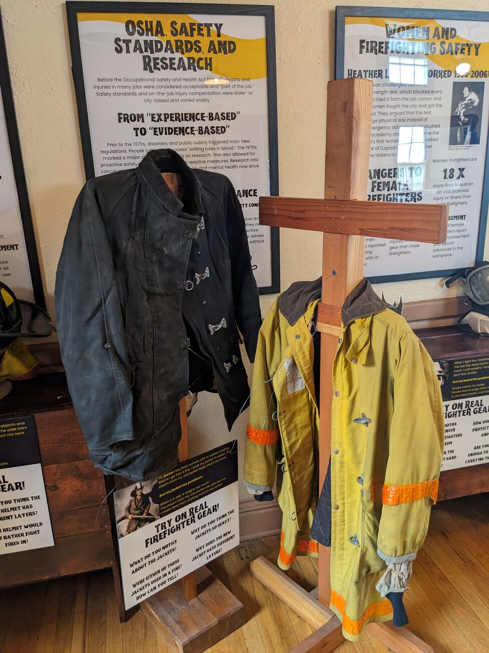 Installation photo: a historic canvas fire coat and a modern turnout coat hang side by side on a handmade wooden rack, mounted in front of the adult interpretive panels.